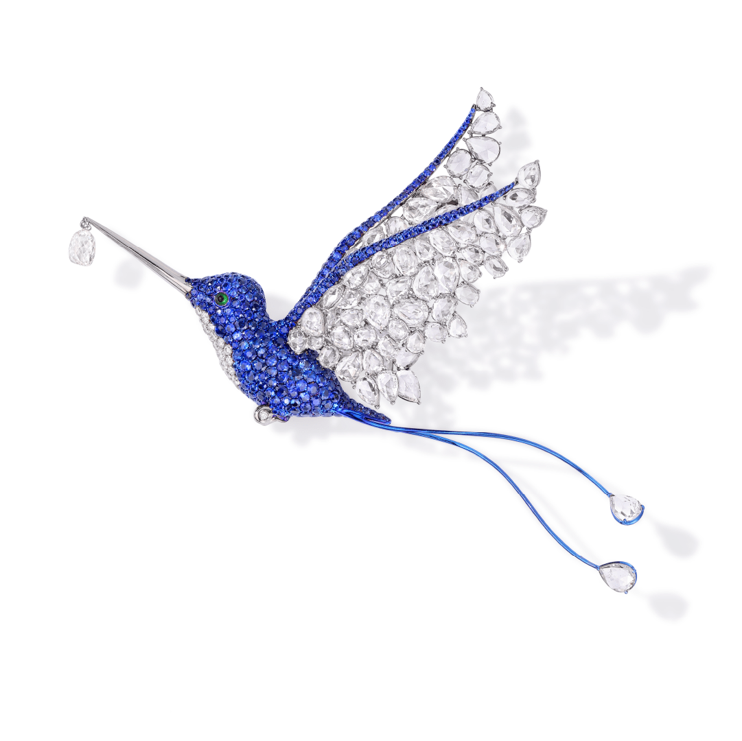 Questa spilla realizzata da Osi Vitoria Jewelry raffigura un colibrì in volo, con un ciondolo di diamanti che ricorda una radiosa goccia di rugiada nel becco. Le ali e la coda scintillano grazie ai diamanti, creando un sorprendente contrasto con il corpo incastonato di zaffiri