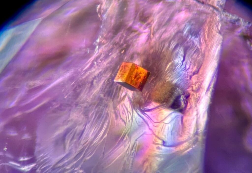 Foto  11: Cubo di pirite in ametista canadese. Un cubo perfetto di pirite sembra galleggiare in questa ametista di Thunder Bay, Canada. L’uso di polarizzatori incrociati esalta l’iridescenza causata dalle fratture all’interno della pietra. Campo visivo circa 2,0 mm