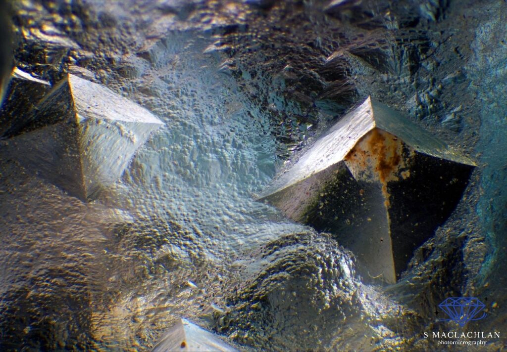Foto 14: Caratteristiche di crescita della fluorite blu dalla Colombia. Si tratta di una sovrapposizione di 52 immagini in stacking scattate con uno zoom di 120x. A confondervi un po’ le idee è il fatto che si tratta di incisioni concave e non di forme piramidali convesse come sembrano! È inoltre possibile notare alcune tracce di ferro nella cavità a destra